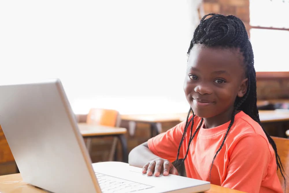 A child smiles while using a laptop in a classroom.