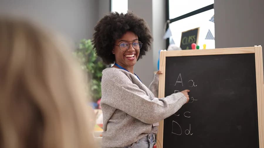 A smiling teacher points to letters on a chalkboard.