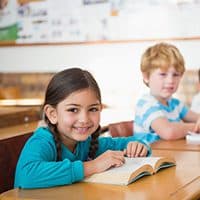 A smiling student reading a book at a classroom desk.