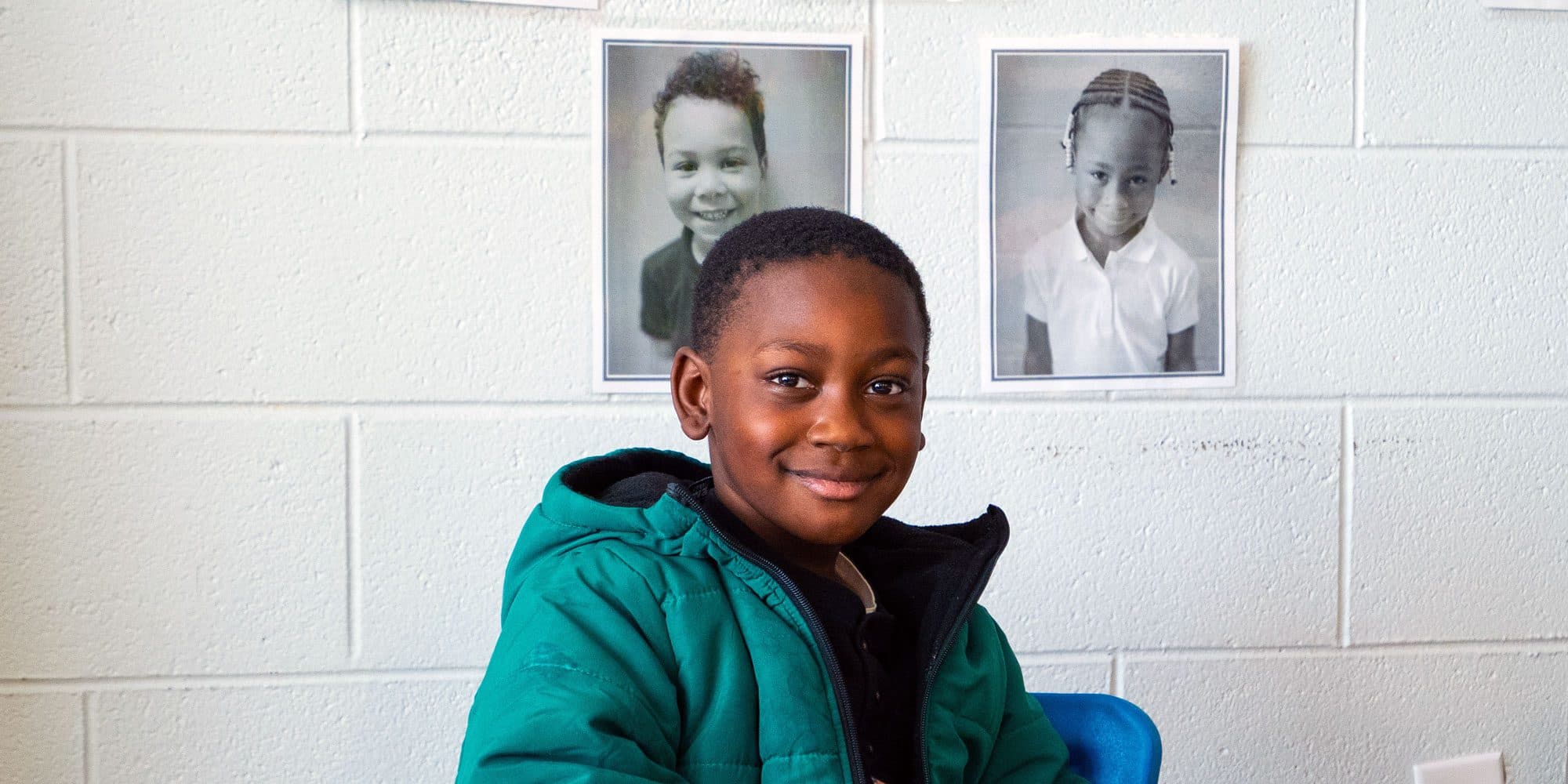 Smiling student in a green jacket in front of a wall of student portraits.