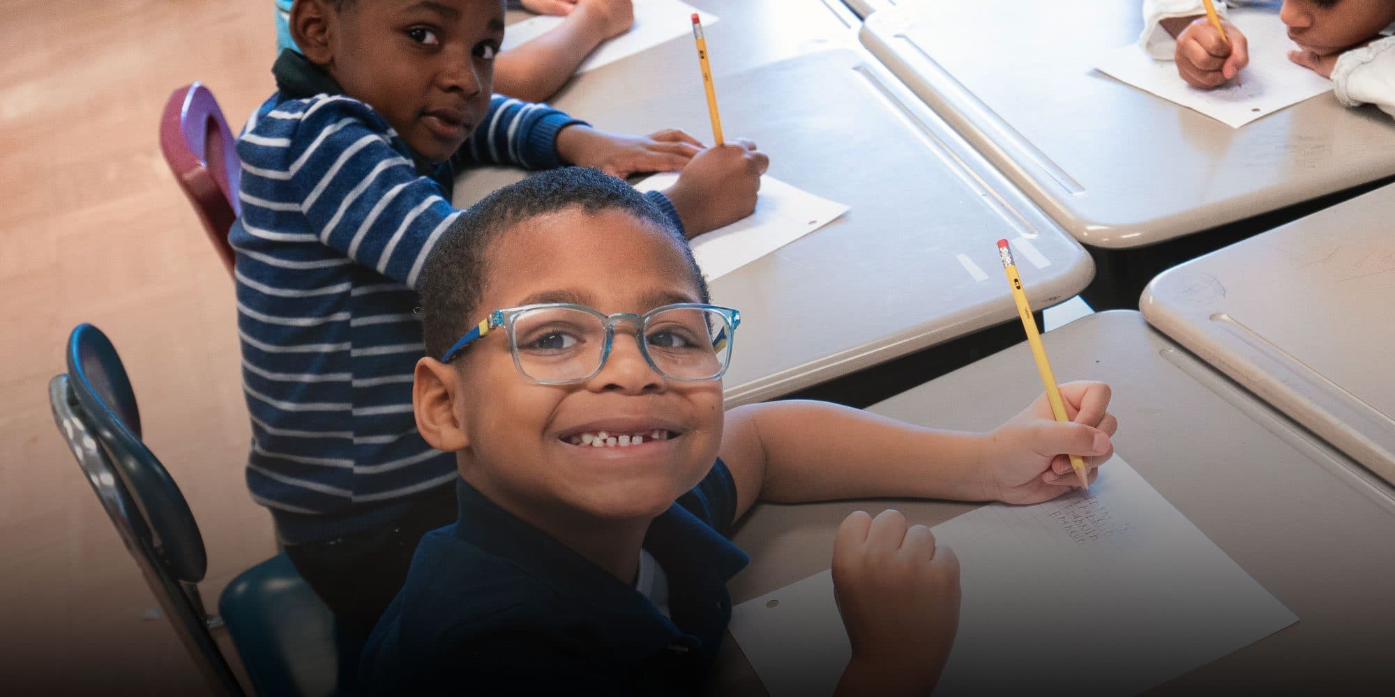 Two students writing at their desks, one smiling at the camera.
