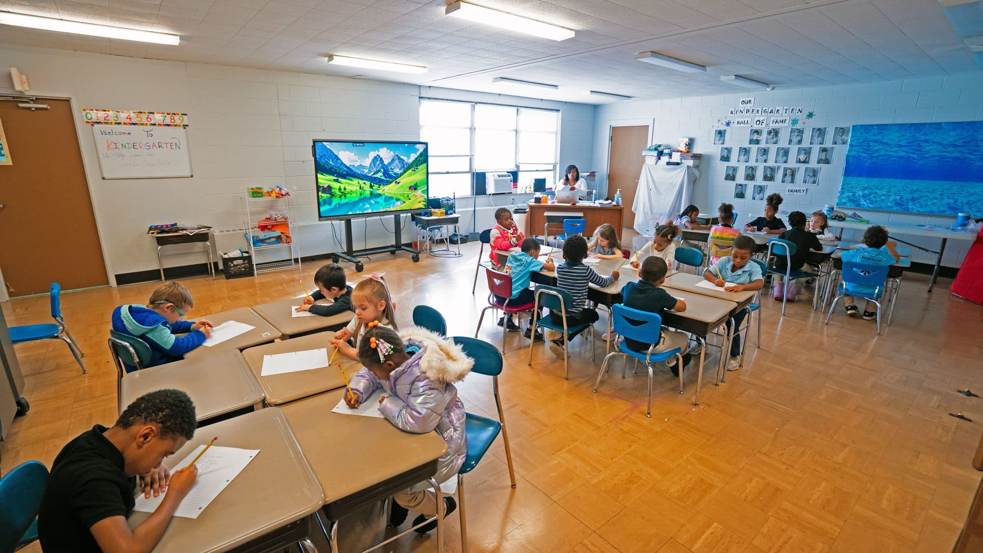 Kindergarten students writing at their desks while a teacher works nearby.