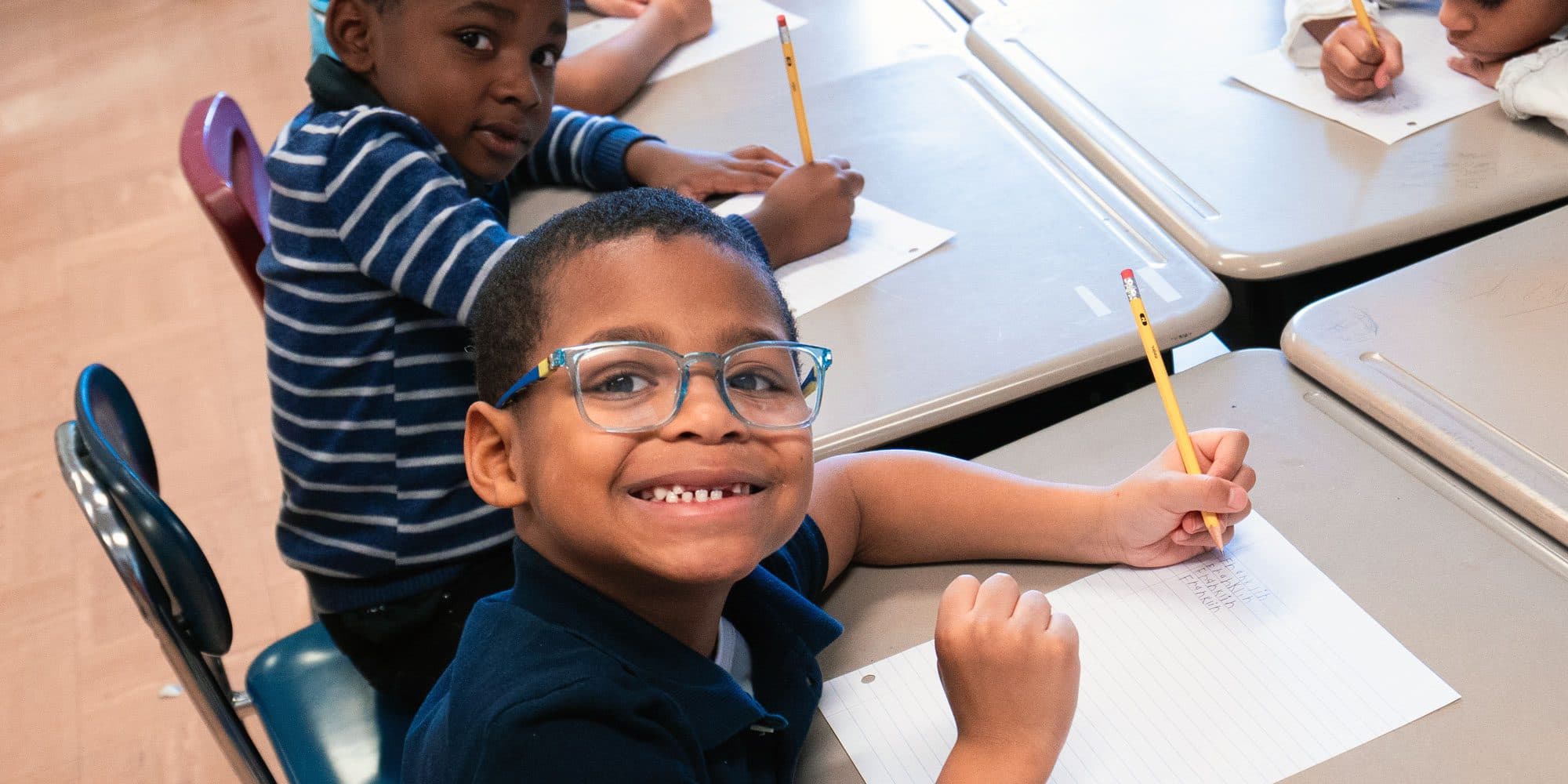 Two students writing at their desks, one smiling at the camera.