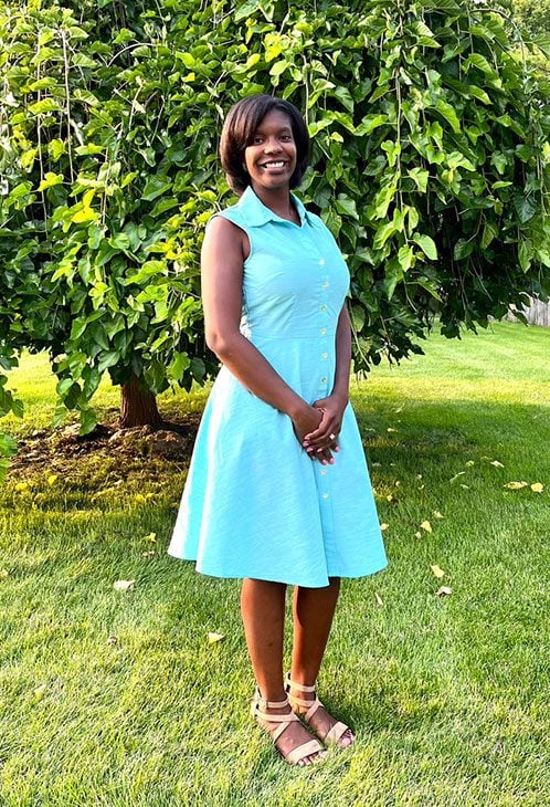 Smiling adult in a light blue dress standing outdoors.